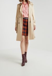 Kvinna iklädd beige trenchcoat, rosa blus, orange och marinblå rutiga kjol, och vinröda ankelstövlar, stående framför en enfärgad vit bakgrund.