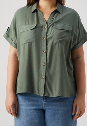 Vrouw draagt een loszittend olijfgroen overhemd met korte mouwen en knopen, met twee borstzakken, gecombineerd met een blauwe spijkerbroek.