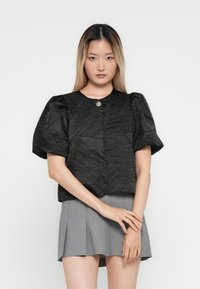 Noor noor, kellel on pikad ombre juuksed, kannab tekstuuriga musta puff-sleeve särk, millel on üks nupp, ja halli voltimata seelikut.