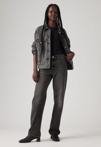 Grijze denimjack over een zwarte top, gecombineerd met taperende grijze jeans en zwarte enkelboots met hak. Model staat voor een lichte achtergrond.