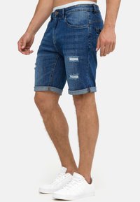 Hombre que lleva puestos unos pantalones cortos de mezclilla azul arremangados, con parches desgastados y zapatillas blancas, de pie contra un fondo liso.
