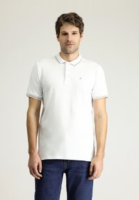 Biała koszulka polo z trzema guziczkami, złotymi akcentami przy kołnierzyku i rękawach, w połączeniu z ciemnoniebieskimi dżinsami. Gładka bawełniana tekstura.