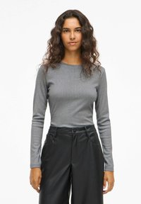 Donna con capelli ondulati e scuri, vestita con un top grigio aderente a maniche lunghe e pantaloni neri a gamba larga a vita alta, in piedi contro uno sfondo bianco.