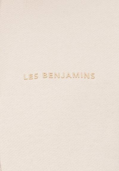 Beidge teksturēts audums ar izsistā zīmola nosaukumu "LES BENJAMINS" nedaudz tumšākā tonī, centrēts apakšā.