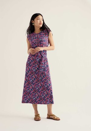 Woman standing wearing sleeveless purple floral midi dress with side knot and tan strappy flat sandals against plain background.