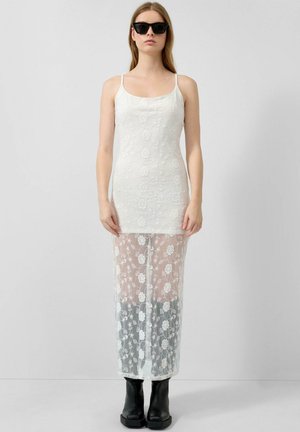 Femme portant une robe blanche en dentelle florale transparente sur un short, des bottes noires et des lunettes de soleil noires, debout devant un fond clair uni.