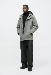 Šedá nepremokavá bunda s kapucňou, zapínaná na gombíky vpredu, s vreckami na ruky, v kombinácii s voľnými čiernymi cargo nohavicami a čiernymi teniskami.