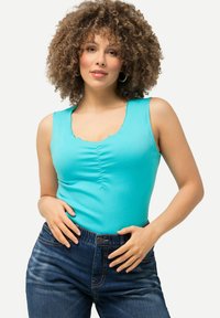 Bodysuit sin mangas en color turquesa con escote redondeado y detalle fruncido en el frente, combinado con jeans de mezclilla oscuros y cierre de botón.