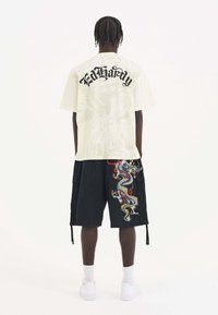 Světle béžové tričko s logem "Ed Hardy" a decentní grafikou na zadní straně; spárované s černými cargo šortkami s barevným výšivkem draka.