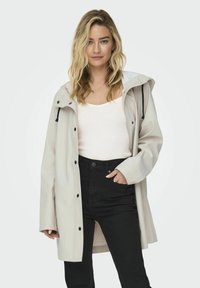 Veste imperméable beige clair avec capuche, dotée de boutons-pression noirs et d'un cordon de serrage, portée par-dessus un débardeur blanc et un pantalon noir.