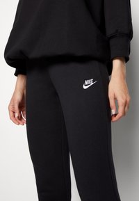 Juodi Nike sportiniai kelniai, pasižymintys prigludusia dizaino forma, minkšta medvilnine medžiaga ir ryškiu baltu Nike logotipu kairiajame šlaunyje.