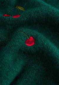 Green knitted fabric with a soft texture, featuring red embroidered cherries and a leaf, creating a subtle pattern.