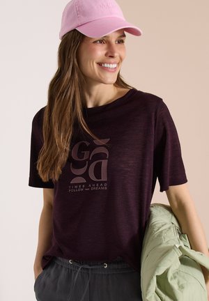 Smilende kvinde iført en pink kasket, mørkelilla grafisk t-shirt med teksten "Times Ahead Follow Your Dreams," mørke bukser, holder en lysegrøn jakke.