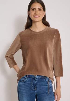 Kvinde smilende med langt mørkt hår iført en brun kordfløjls long-sleeve top og blå denim jeans, stående mod en ensfarvet baggrund.