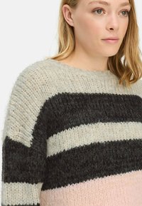 Vrouw in een chunky gebreide trui met horizontale strepen in crème, zwart en lichtroze, getoond van schouders tot kin.