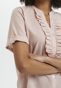 Camicia a maniche corte, con bottoni, di un morbido rosa con righe bianche, caratterizzata da una pettorina arricciata e maniche arrotolate fissate con bottoni.