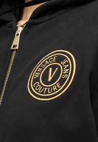 Μαύρο τζάκετ με φερμουάρ και χρυστό κυκλικό λογότυπο Versace Jeans Couture κεντημένο στο στήθος κοντά στο φερμουάρ.