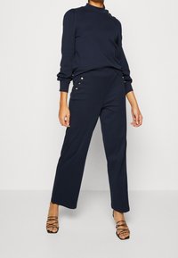 Navy sweatshirt dengan leher tinggi dan aksen kancing emas, dipadukan dengan celana navy lebar yang memiliki detail kancing emas di sisi.