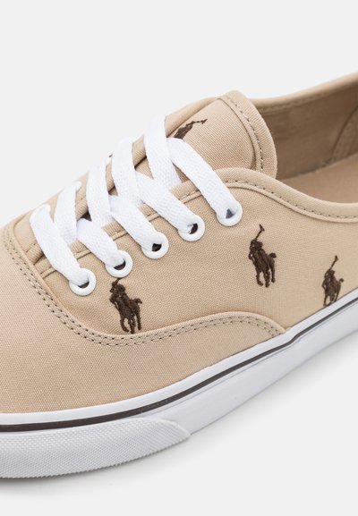 Sapatilha de tela bege com atacadores brancos, apresentando os logótipos bordados em castanho e uma sola de borracha branca com uma faixa escura.