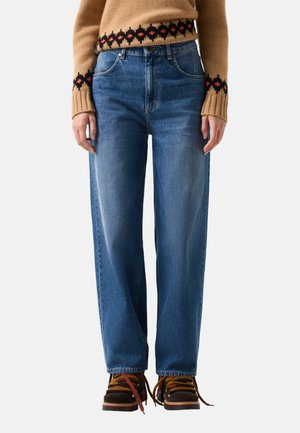 Person iført højttaljede, lige blå jeans, brun sweater med sort og rødt mønster samt brune vandrestøvler med røde snørebånd.