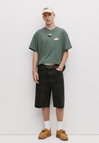 Olivgrünes T-Shirt mit einem kleinen orangefarbenen Grafik, schwarze karierte Shorts, beige Kappe und braune Schuhe. Silberne Accessoires und ein schwarzer Gürtel.