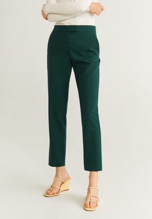 Frau trägt dunkelgrüne Hose mit geradem Bein, kombiniert mit beigen Keilsandalen mit Riemchen, steht mit verschränkten Armen.