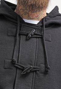 Sweat-shirt à capuche noir avec une texture lisse, doté d'une fermeture à bascule et de cordons ajustables. Design minimaliste avec des surpiqûres décoratives.