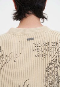 Beige gerippter Pullover mit strukturiertem Strick, bedruckten schwarzen Grafiken und einem kleinen Metalltag am hinteren Ausschnitt.