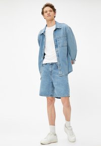 Chaqueta de mezclilla y pantalones cortos en un lavado azul claro, combinados con una camiseta blanca. Zapatillas blancas completan el atuendo casual.