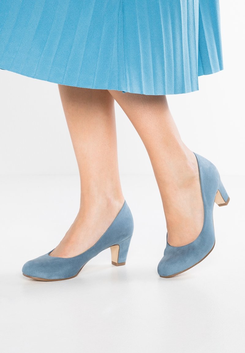 Femme portant des escarpins en daim bleu à talon moyen et une jupe plissée assortie, debout sur un sol blanc.