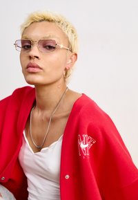 Red cardigan with a Nike logo, white t-shirt underneath, gold-accented glasses, short blonde hair, multiple ear piercings, heart tattoo on neck.