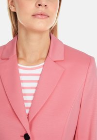 Light pink blazer featuring a tailored collar, single button closure, layered over a white and pink striped top. Smooth fabric texture.