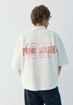 Młody mężczyzna z krótkimi kręconymi włosami, ubrany w luźną białą koszulę z krótkim rękawem z napisem "1991 PRIME SEASON" na plecach w kolorze czerwonym.