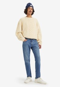 Pull en tricot à grosses mailles crème, jeans bleus et baskets blanches. Stylisé avec un bonnet bleu marine. Coupe décontractée et matériaux texturés.