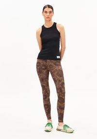 Een jonge vrouw staat zelfverzekerd, gekleed in een zwarte tanktop en bruine geprinte leggings, gecombineerd met sneakers met groene veters.
