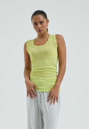 Femme aux cheveux tirés en arrière, portant un haut sans manches vert clair et un pantalon ample gris clair, debout avec les mains près des hanches.