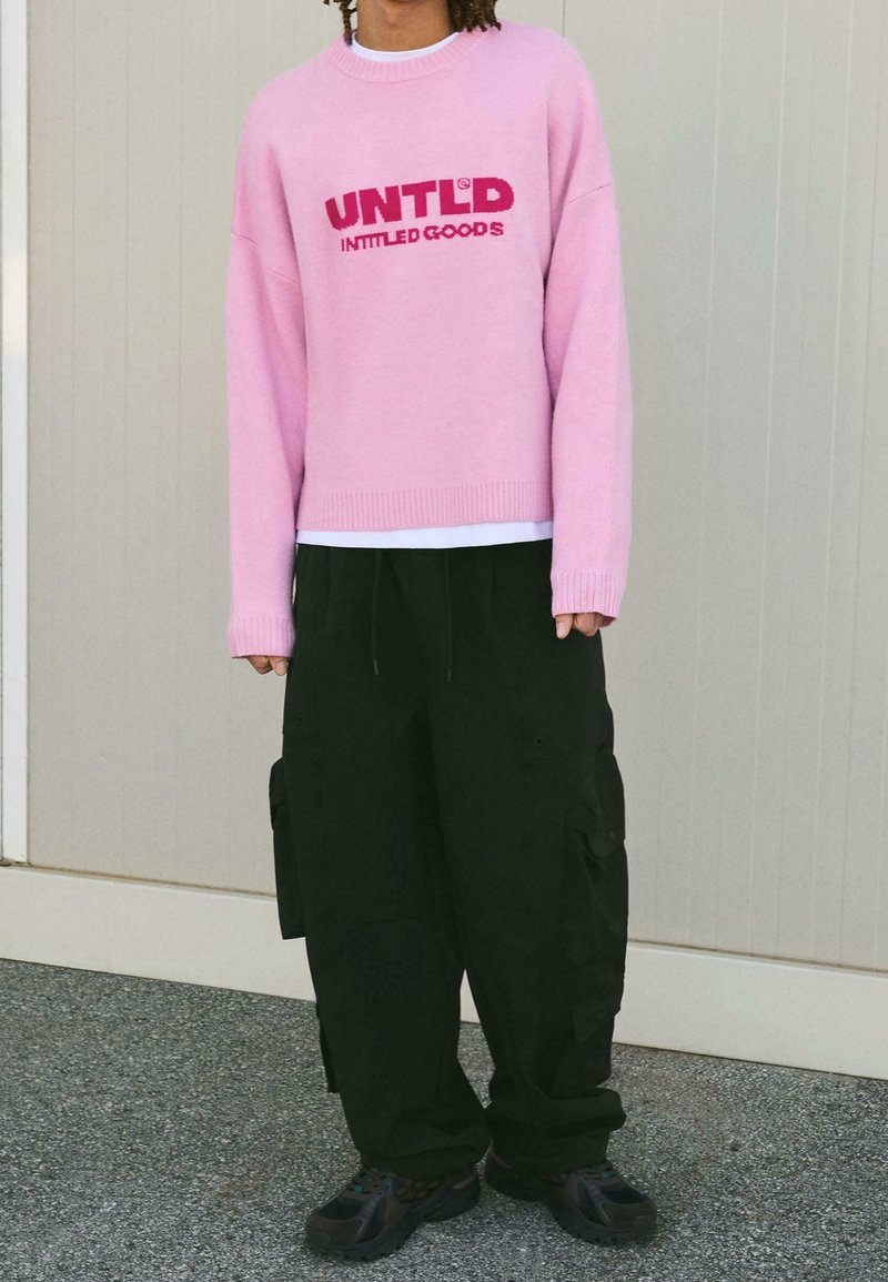 Personne portant un pull rose clair avec un texte de logo rouge, un pantalon cargo noir et des baskets noires, debout contre un mur beige.