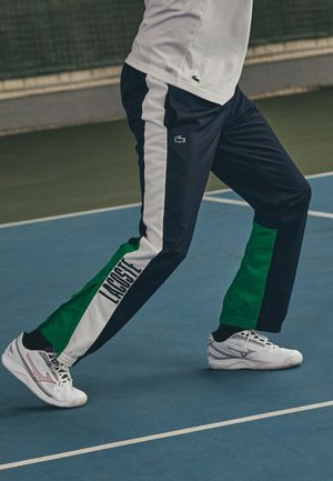 Person in marineblauen, weißen und grünen Lacoste-Trainingshosen und weißen Turnschuhen, die auf einem blauen Tennisplatz ausfällt.