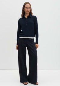 Donna in piedi con polo nera a maniche lunghe abbottonata, pantaloni neri a gamba larga e tacchi neri aperti davanti, su sfondo bianco uniforme.