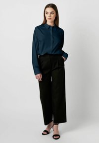 Dunkelbluse aus Tealfarbe mit Knopfleiste und langen Ärmeln. Kombiniert mit hochtaillierten schwarzen, weit geschnittenen Hosen und schwarzen Riemchen-Heels.