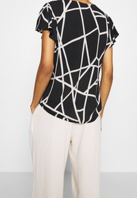 Černá blůza s lehkým béžovým geometrickým vzorem, krátké volány na rukávech, volný střih a zakřivený lem, spárována se světle zbarvenými strukturovanými kalhotami.