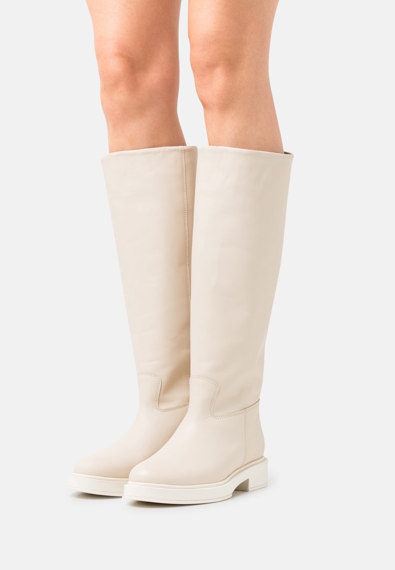 Knee-high vaalean kreemivärvi nahast saapad, mis on kantud paljaste jalgadega, ümardatud varvaste ja madalate plokk-kontsadega, valge tausta peal.