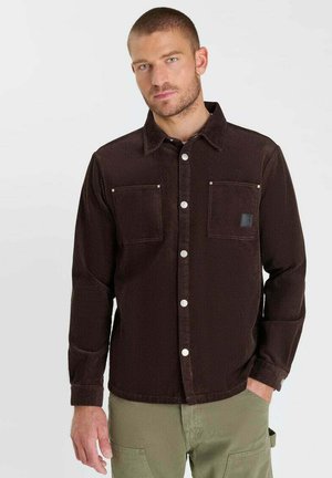 Uomo con capelli corti che indossa una camicia di velluto a coste marrone scuro con bottoni e pantaloni verde chiaro, in piedi con una mano in tasca.