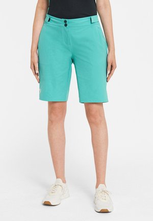 Frau trägt türkisfarbene kniehohe Shorts, schwarzes Oberteil und weiße Turnschuhe, stehend vor schlichtem, hellem Hintergrund.