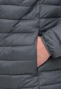 Ruka vložená do bočného vrecka tmavosivej prešitej páperovej bundy so zreteľnými horizontálnymi švami a elastickým lemom.