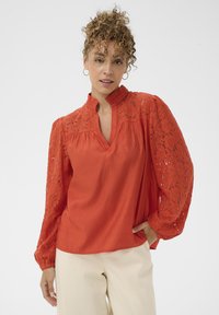 Blouse rouge à col montant, avec des manches en dentelle et un corps texturé. Manches longues et amples, coupe décontractée.