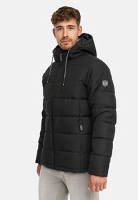 Junger Mann mit kurzen lockigen Haaren, der eine schwarze gepolsterte Jacke mit Kapuze und hellgraue Hosen trägt und vor einem schlichten Hintergrund steht.