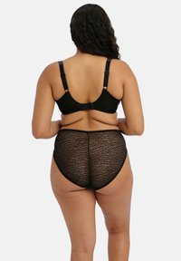 Een plus-size vrouw van achteren afgebeeld, met een zwart verstelbaar bh en hoge doorzichtige panties met luipaardprint, tegen een neutrale achtergrond.