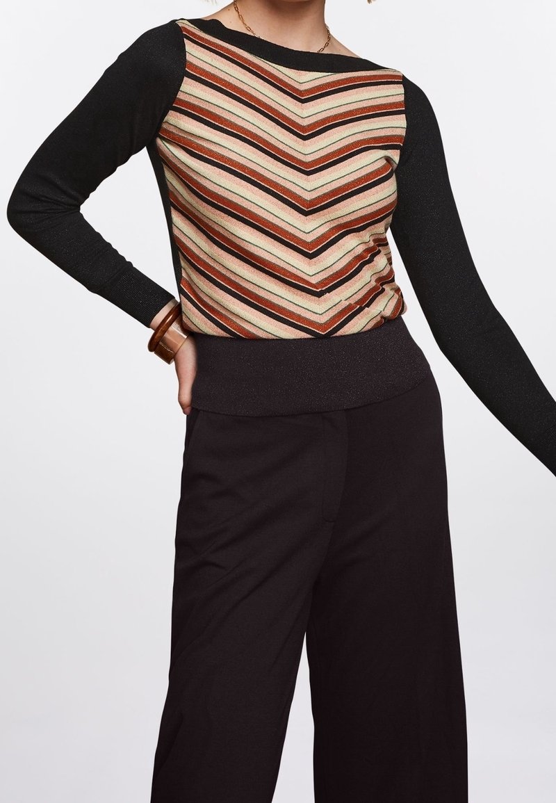 Femme portant un haut à manches longues avec des manches noires et des rayures en chevrons multicolores sur le devant, associé à un pantalon noir à jambes larges.