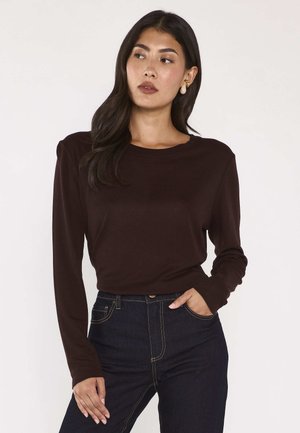 Donna con capelli lunghi e scuri che indossa una maglia a maniche lunghe marrone scuro e jeans blu scuro, guarda di lato con una mano in tasca.